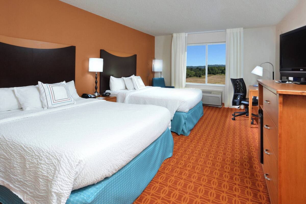 A hotel room with two beds, a desk, a TV, and a large window offering a view of the outdoors.