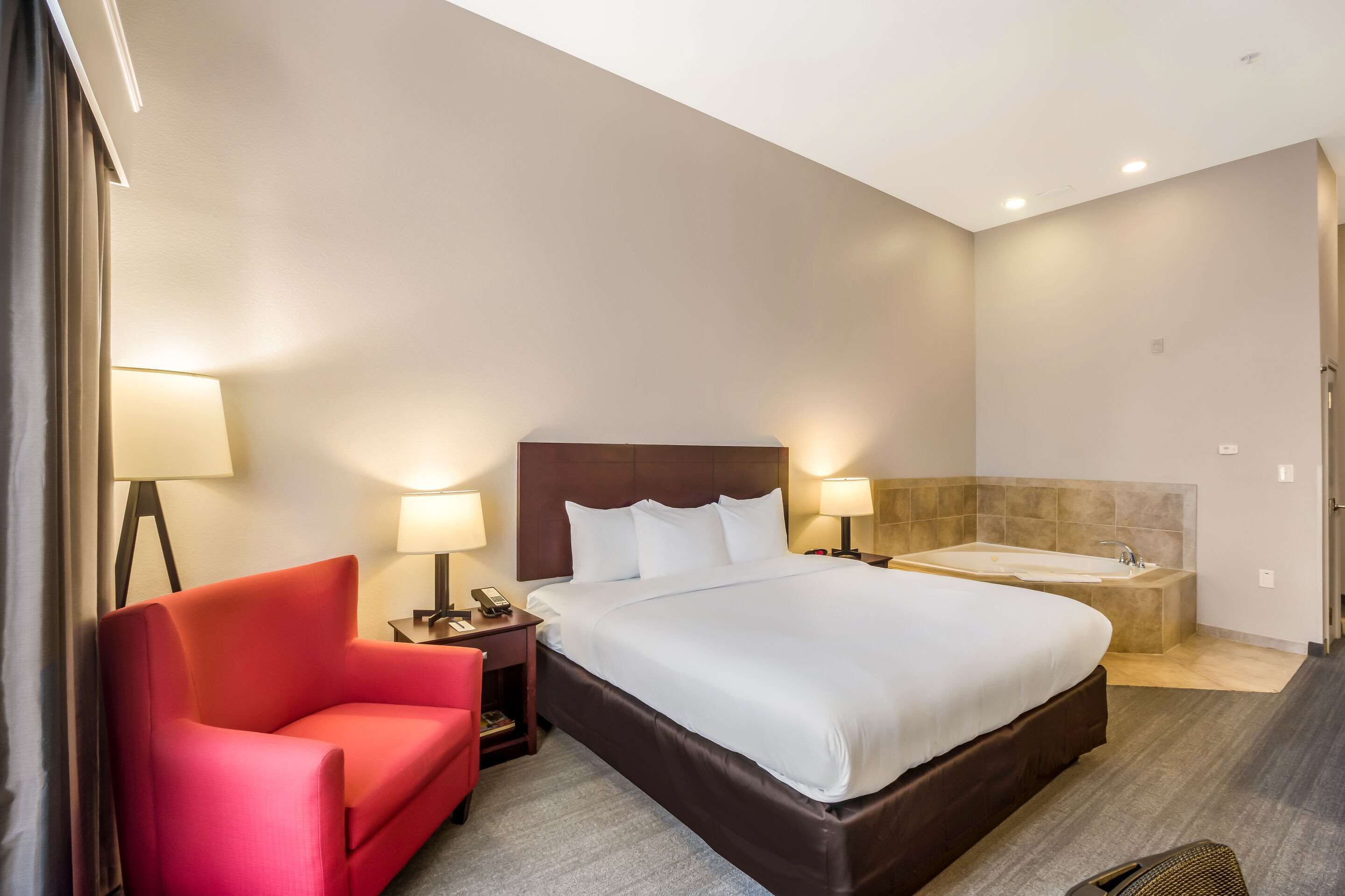 A hotel room with a large bed, a red chair, a nightstand with a lamp, and a bathtub.