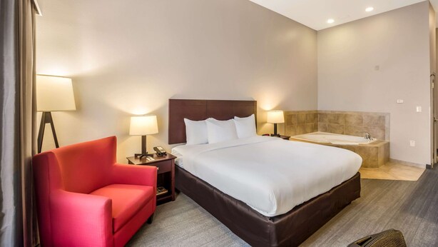 Quarto de hotel com uma cama grande, uma cadeira vermelha, uma mesa de cabeceira com abajur e uma banheira.