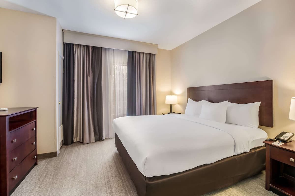 Une chambre d’hôtel avec un grand lit, une table de chevet avec une lampe, une commode et une fenêtre avec des rideaux.