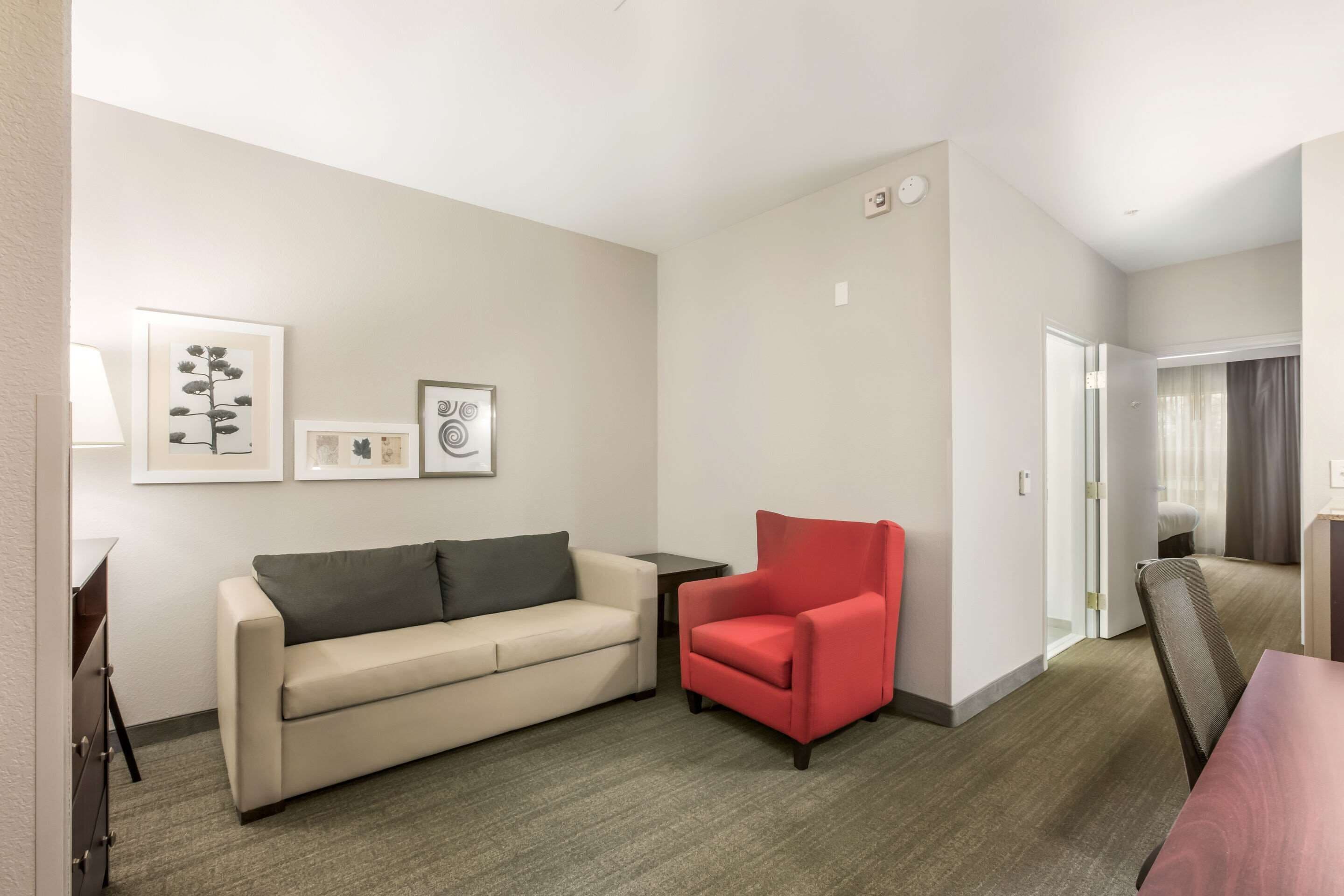 Quarto de hotel com sofá bege, poltrona vermelha, mesinha e obras de arte emolduradas na parede.
