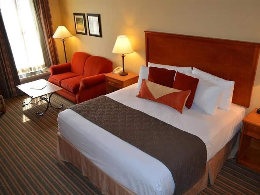 Una habitación de hotel con una cama grande, un sillón rojo, dos lámparas de noche y una mesita con lámpara.