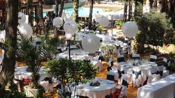 Un área de comedor al aire libre con mesas redondas cubiertas con manteles blancos, rodeada de follaje y árboles.
