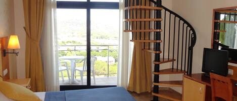 Habitación de hotel con cama, escritorio con computadora, televisor, escalera en espiral y balcón con vistas.