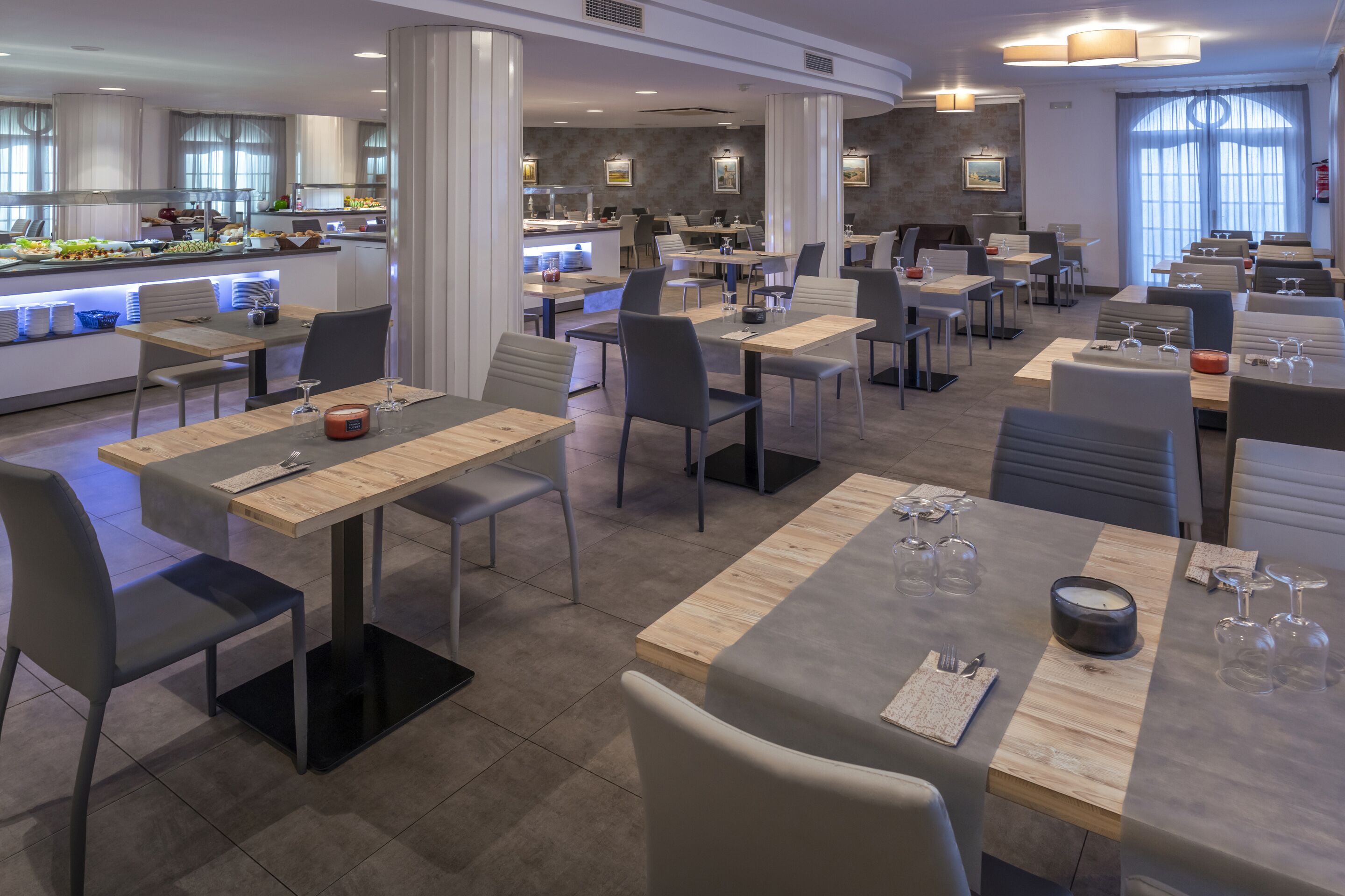 Un restaurante moderno con varias zonas de comedor, incluyendo un bar y un bufé. Las mesas están preparadas con cubiertos y copas de vino.
