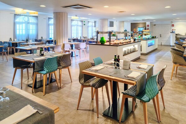 A modern hotel dining area with multiple tables and chairs, a central bar, and a kitchen in the background. - Golden Acqua Salou (Salou)