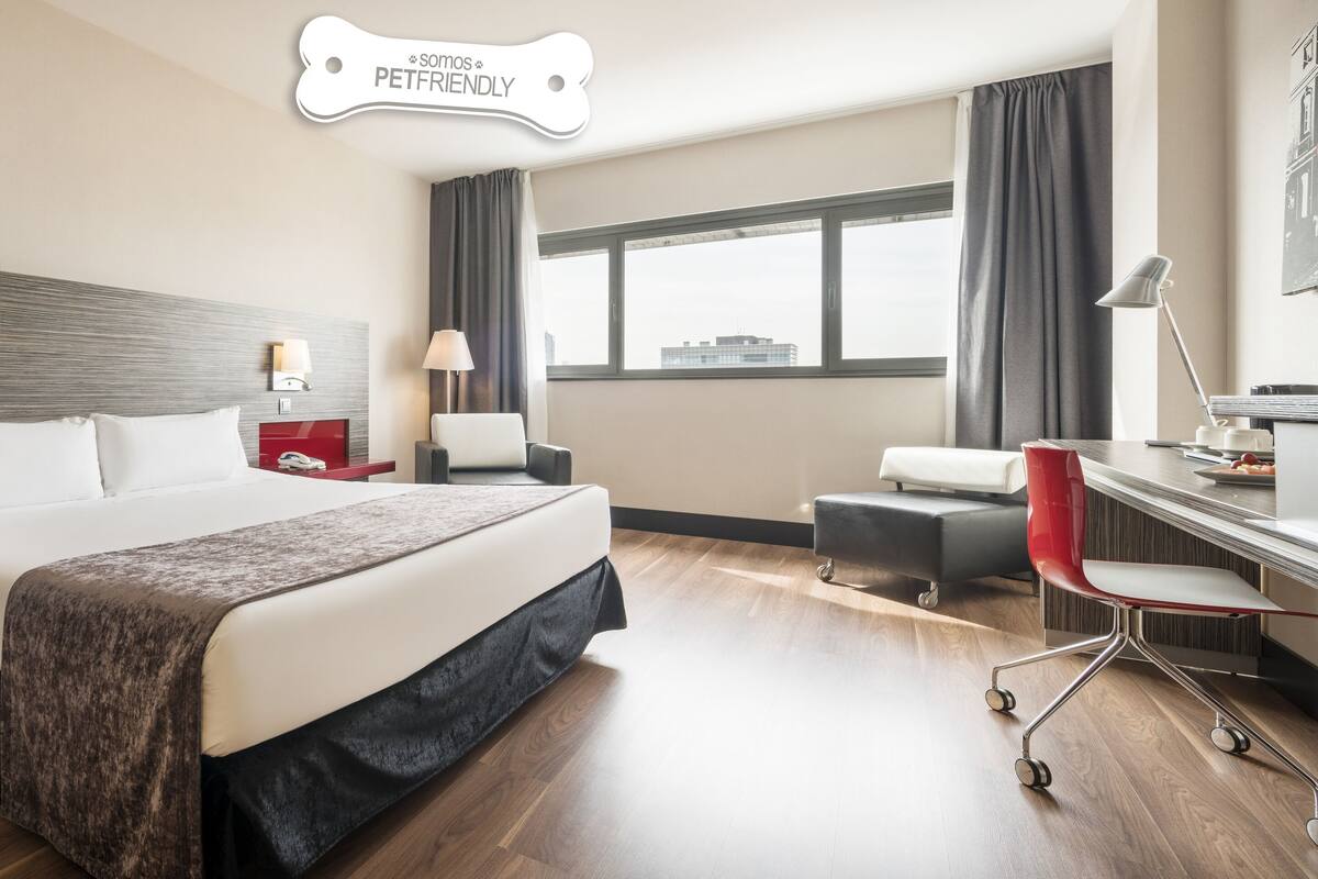 A modern hotel room with a large bed, a desk with a chair, a desk lamp, and a view of the city through the window.