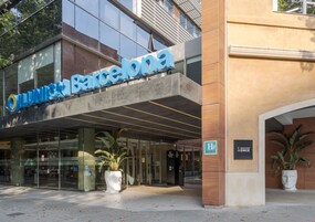Junio Barcelona -hotellin ulkopuoli: lasijulkisivu, suuri ruukkuistutettu kasvi sisäänkäynnin yhteydessä ja oikealla puolella oleva kyltti, joka osoittaa sosiaalisen ryhmän kokoontumispaikan.