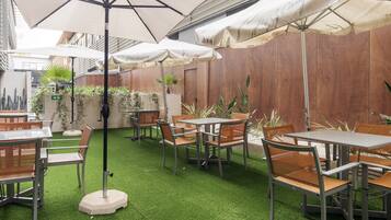 Un patio al aire libre con mesas y sillas, cubierto por sombrillas y rodeado de plantas.