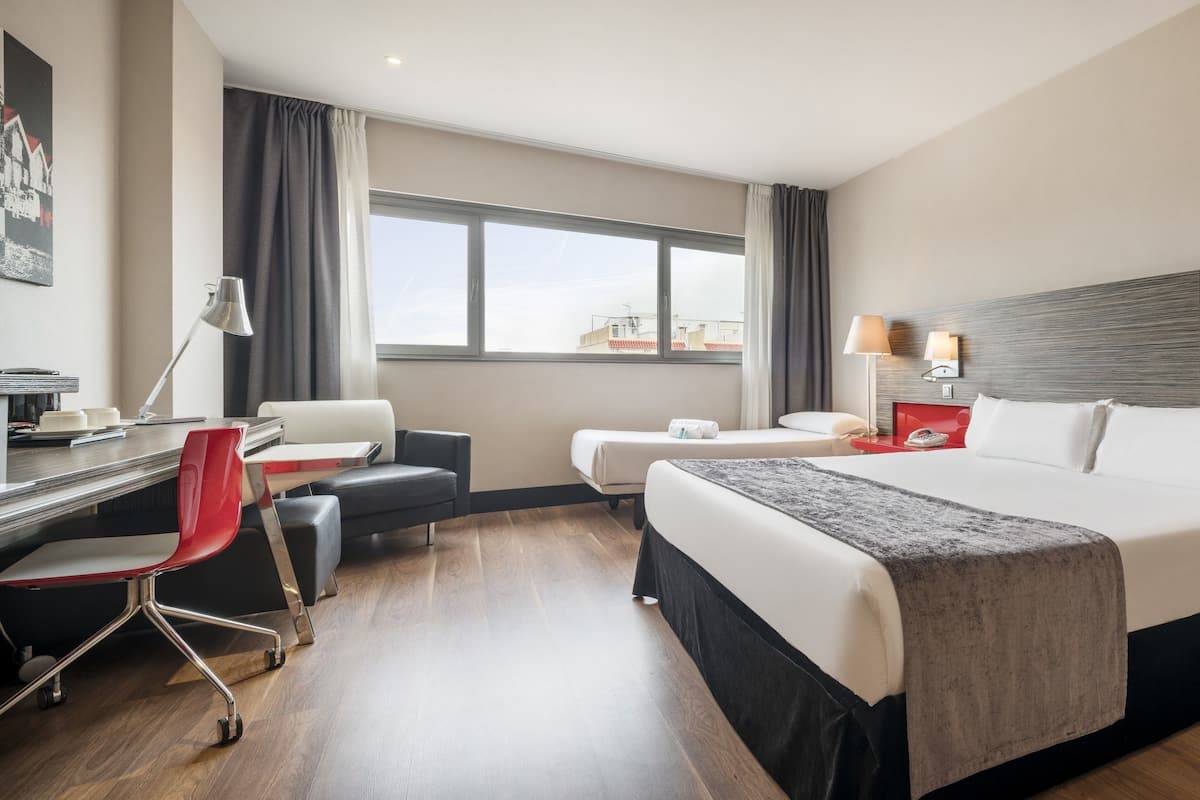 A modern hotel room with a large bed, a desk with a chair, a small table, and a window with curtains.