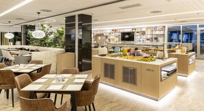 A modern restaurant with a central buffet area, seating with tables and chairs, and a decorative plant arrangement.