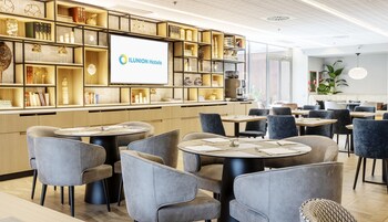 Un moderno vestíbulo de hotel con mesas y sillas redondas, una gran pantalla con el nombre del hotel y una estantería bien organizada con varios objetos decorativos.