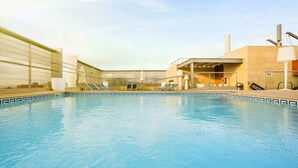Una piscina con sillones de descanso y una zona cubierta, rodeada por un edificio con grandes ventanales y fachada de baldosas.