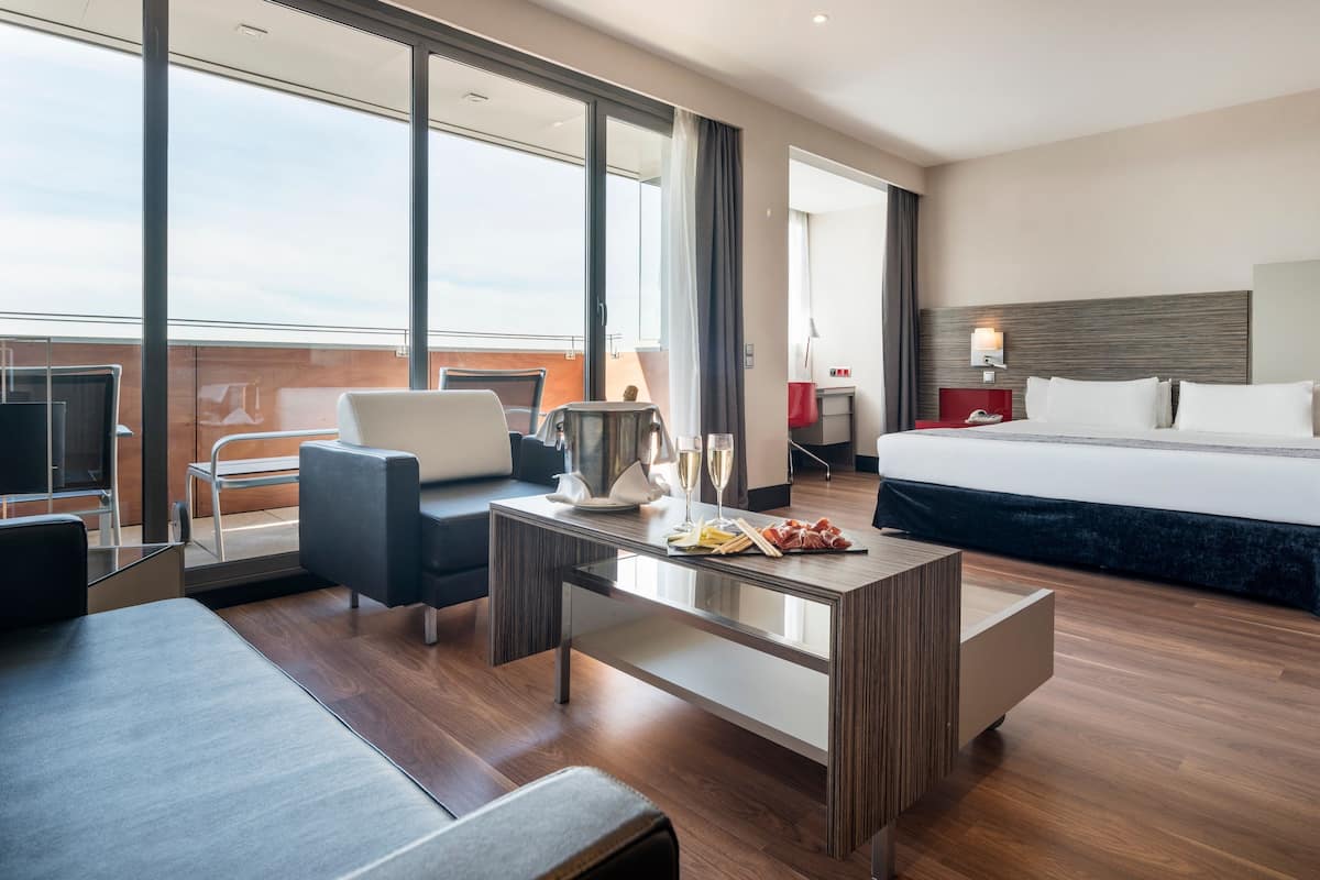 A modern hotel room with a large bed, a desk with a chair, a sofa, and a view of the city through large windows.