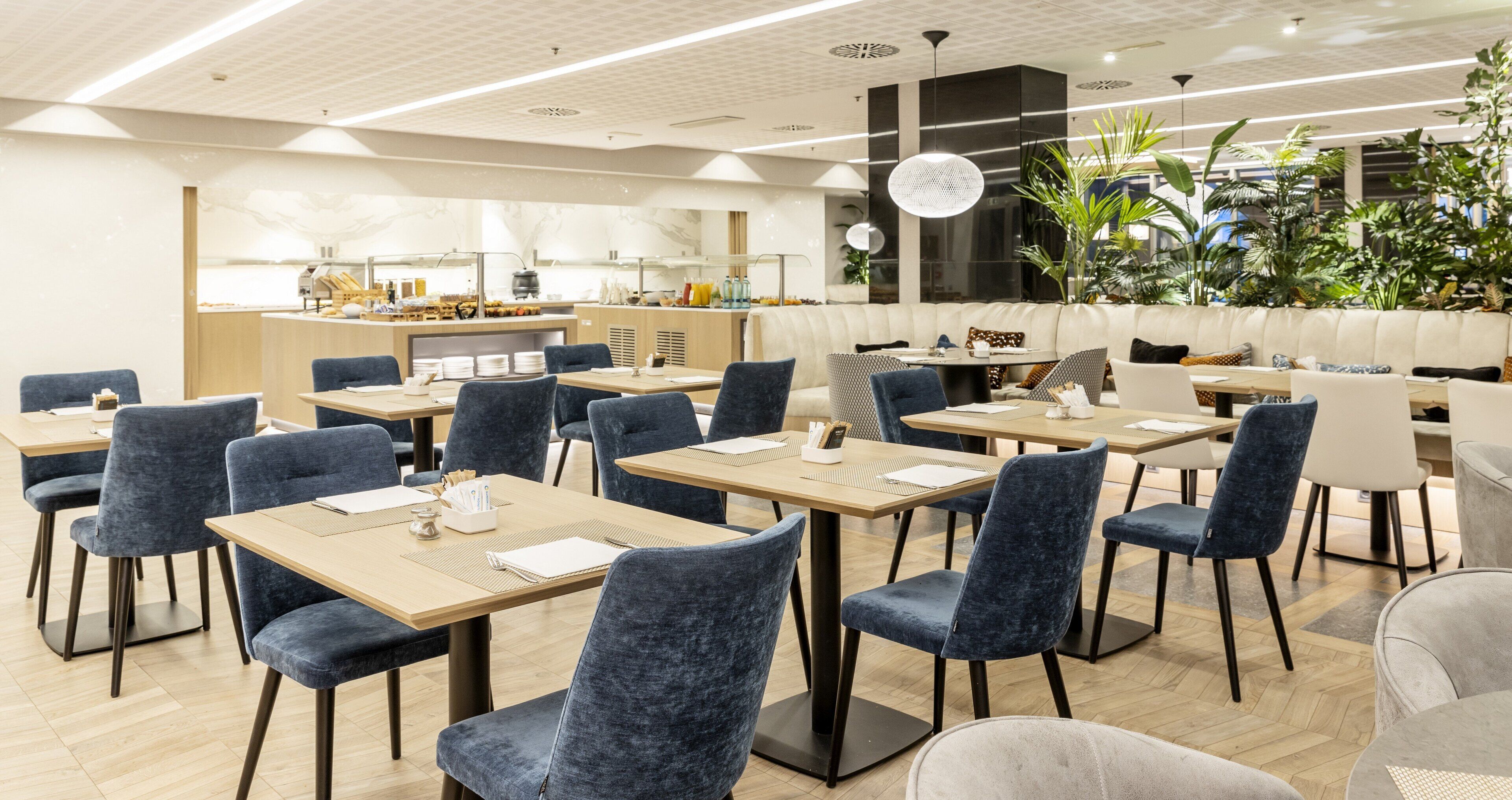 Un restaurante moderno con mesas y sillas, una barra con productos alimenticios y un amplio ventanal con plantas.