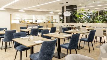 Un restaurante moderno con mesas y sillas, una barra con productos alimenticios y un amplio ventanal con plantas.