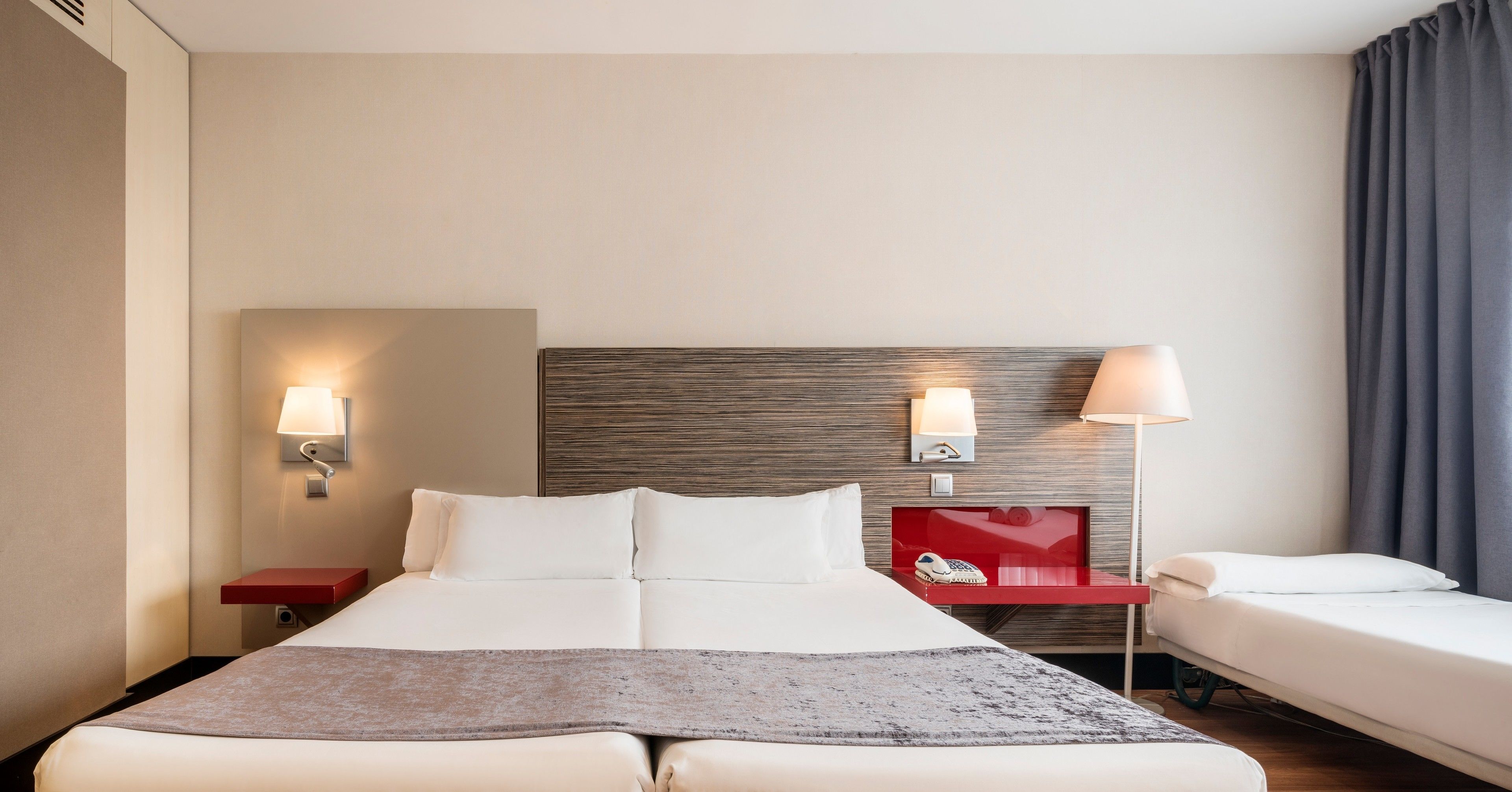 A hotel room with a large bed, two bedside tables, a red nightstand, a headboard, and a window with curtains.