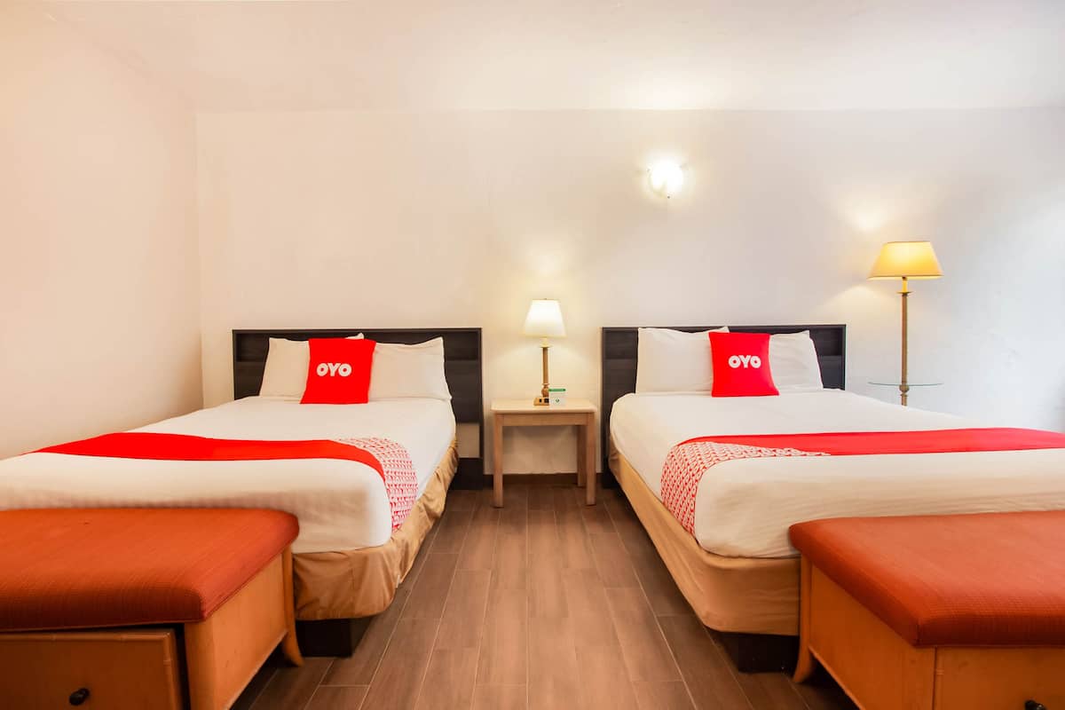 Habitación de hotel con dos camas, cada una con colcha roja y blanca, una mesita de noche con lámpara y suelo de madera.
