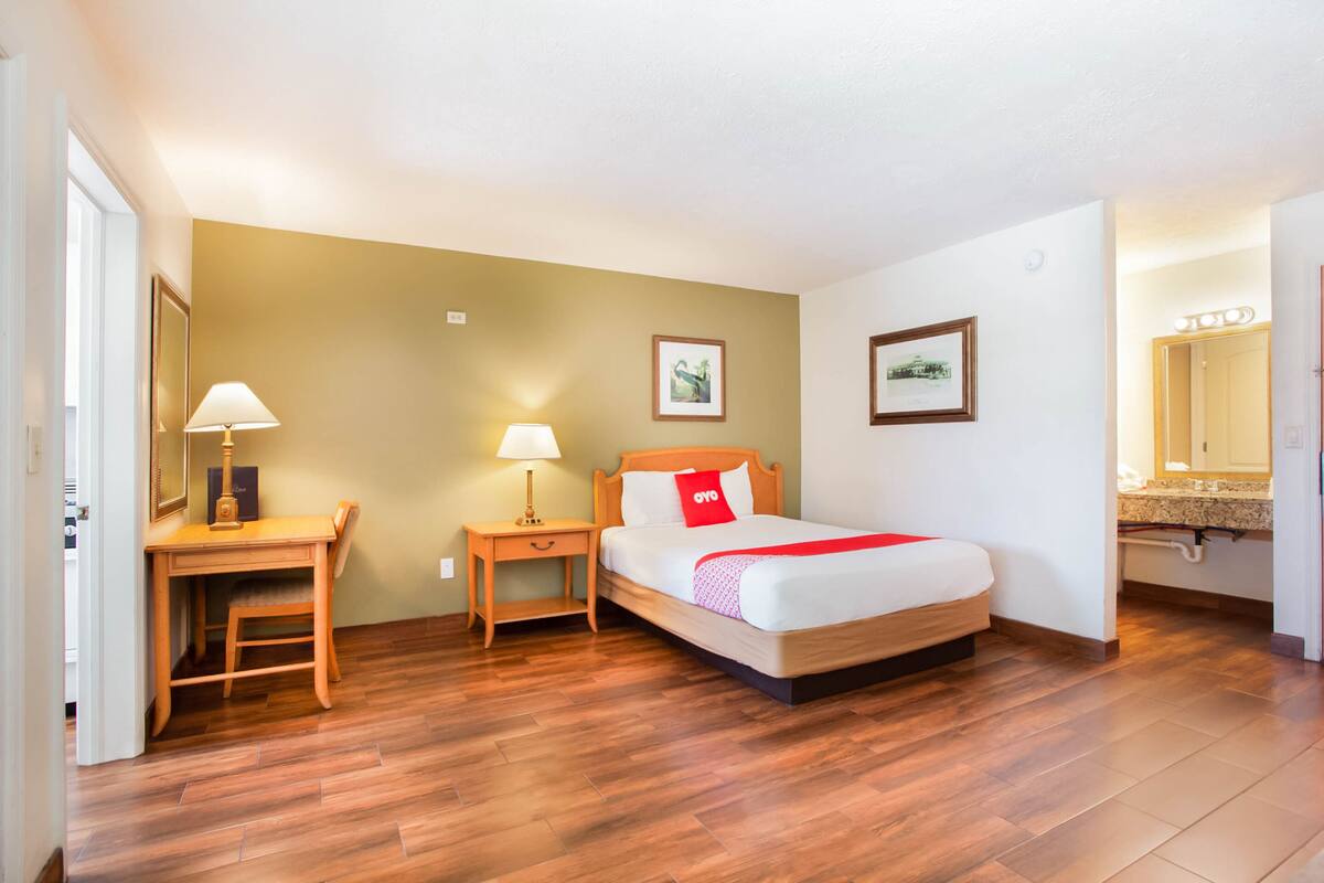 Una habitación de hotel con un escritorio de madera, una cama con un cojín rojo y una mesita de noche con una lámpara.
