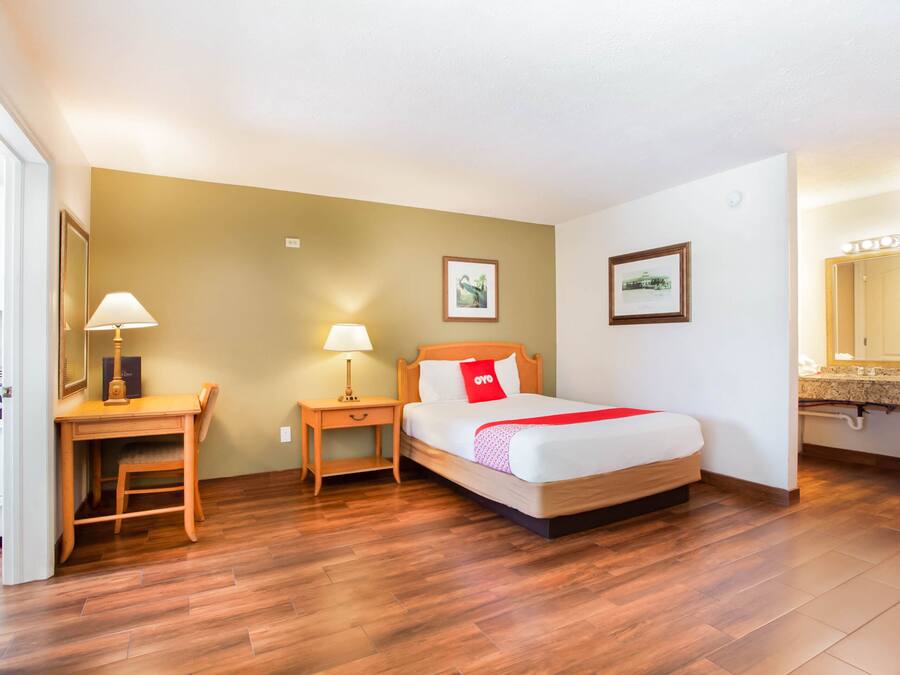 A hotel room with a wooden desk, a bed with a red pillow, and a nightstand with a lamp.