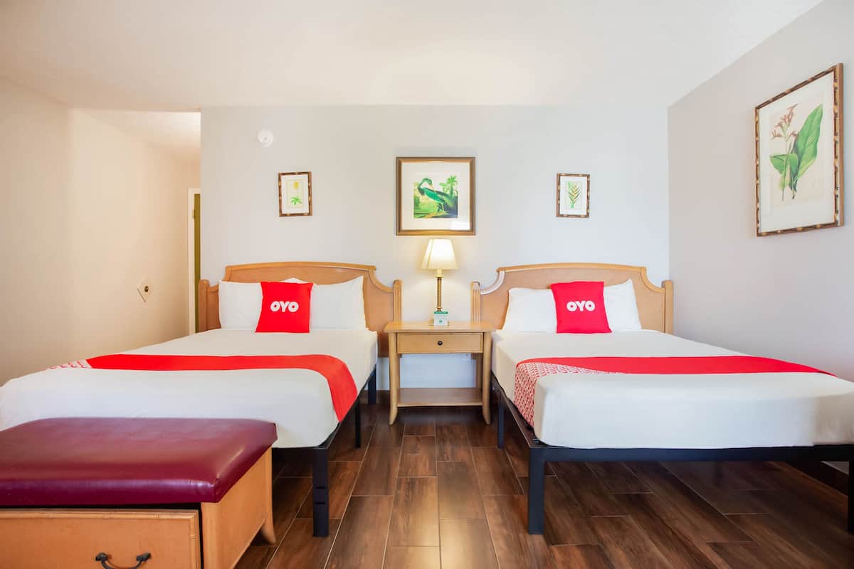 Una habitación de hotel con dos camas individuales, una mesita de noche de madera, una lámpara y un banco.