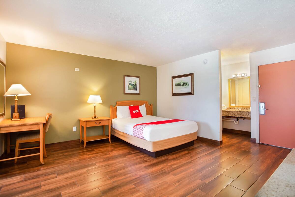 Habitación de hotel con escritorio de madera, cama con almohada roja, mesita de noche con lámpara y baño visible a través de una puerta abierta.