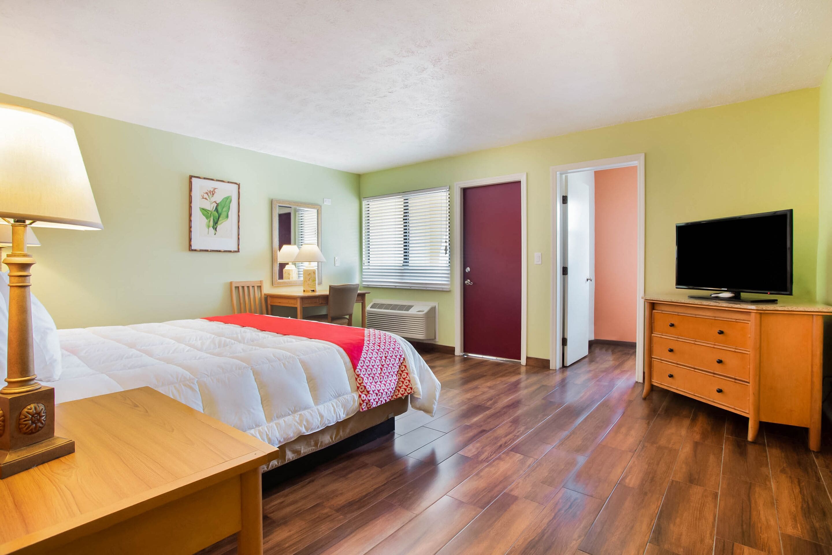 A hotel room with a bed, a desk, a lamp, a television, and a door.