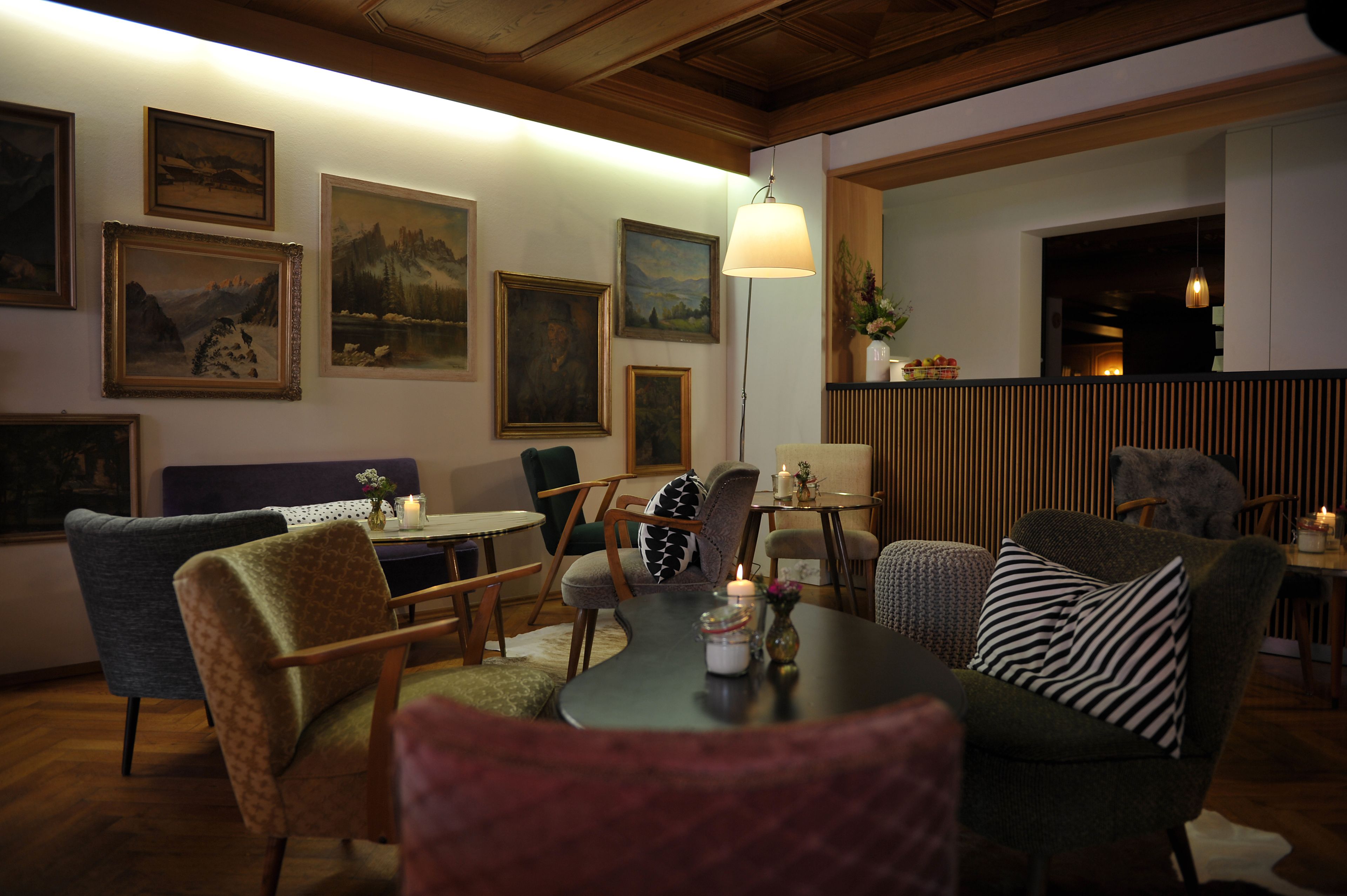 A cozy living room with a wooden ceiling, a round table, and several chairs. There are framed pictures on the walls and a lamp on a side table.