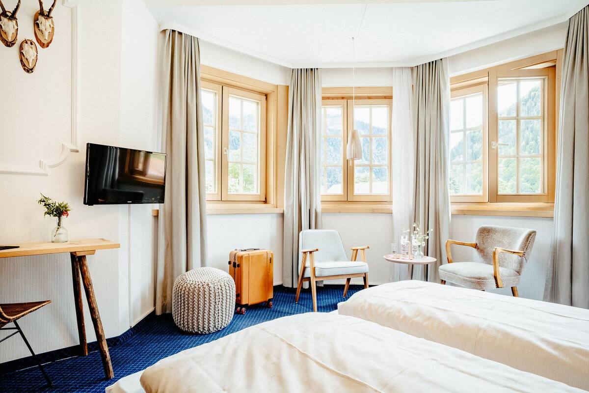 Ein Hotelzimmer mit zwei Betten, einem Fernseher, einem Schreibtisch, einem Sessel, einem kleinen Tisch und großen Fenstern mit Vorhängen.