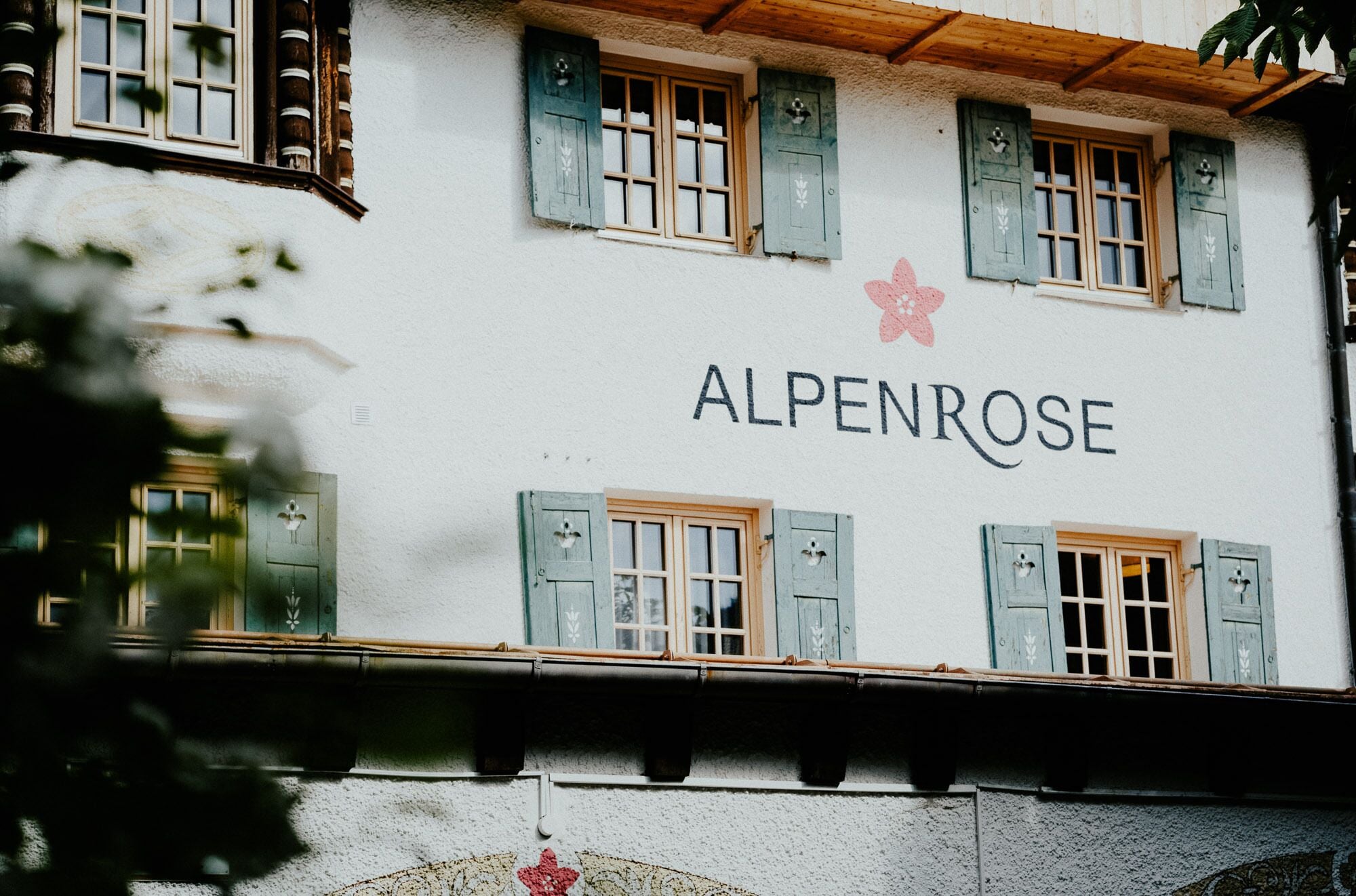 Un edificio con el nombre "ALPENROSE" en su fachada, con contraventanas de madera tradicionales y un estilo arquitectónico rústico y sencillo.