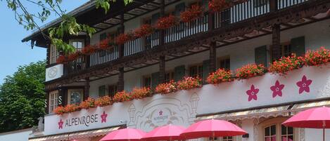Un edificio tradicional con balcones decorados con flores, un letrero que dice "ALPENROSE" y varios paraguas rojos.