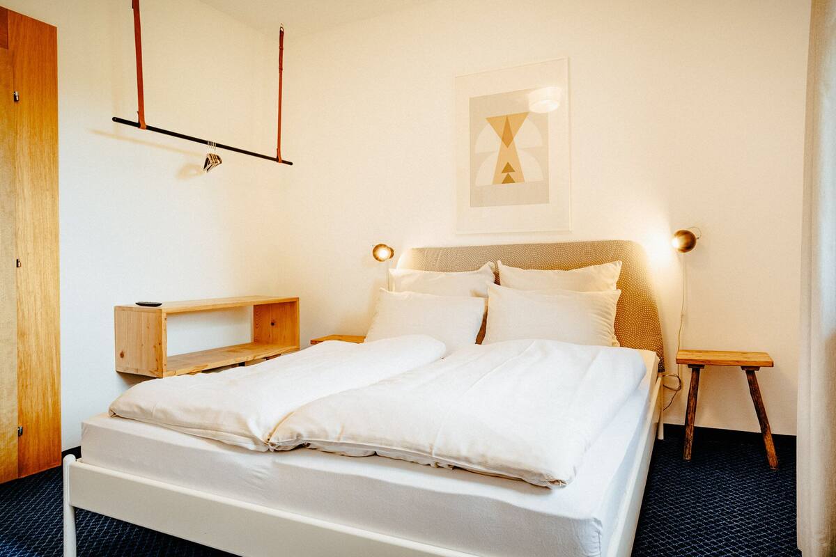 Ein ordentlich bezogenes Bett mit weißen Leinen, ein Nachttisch aus Holz, eine Wandlampe und ein gerahmtes Bild über dem Bett.