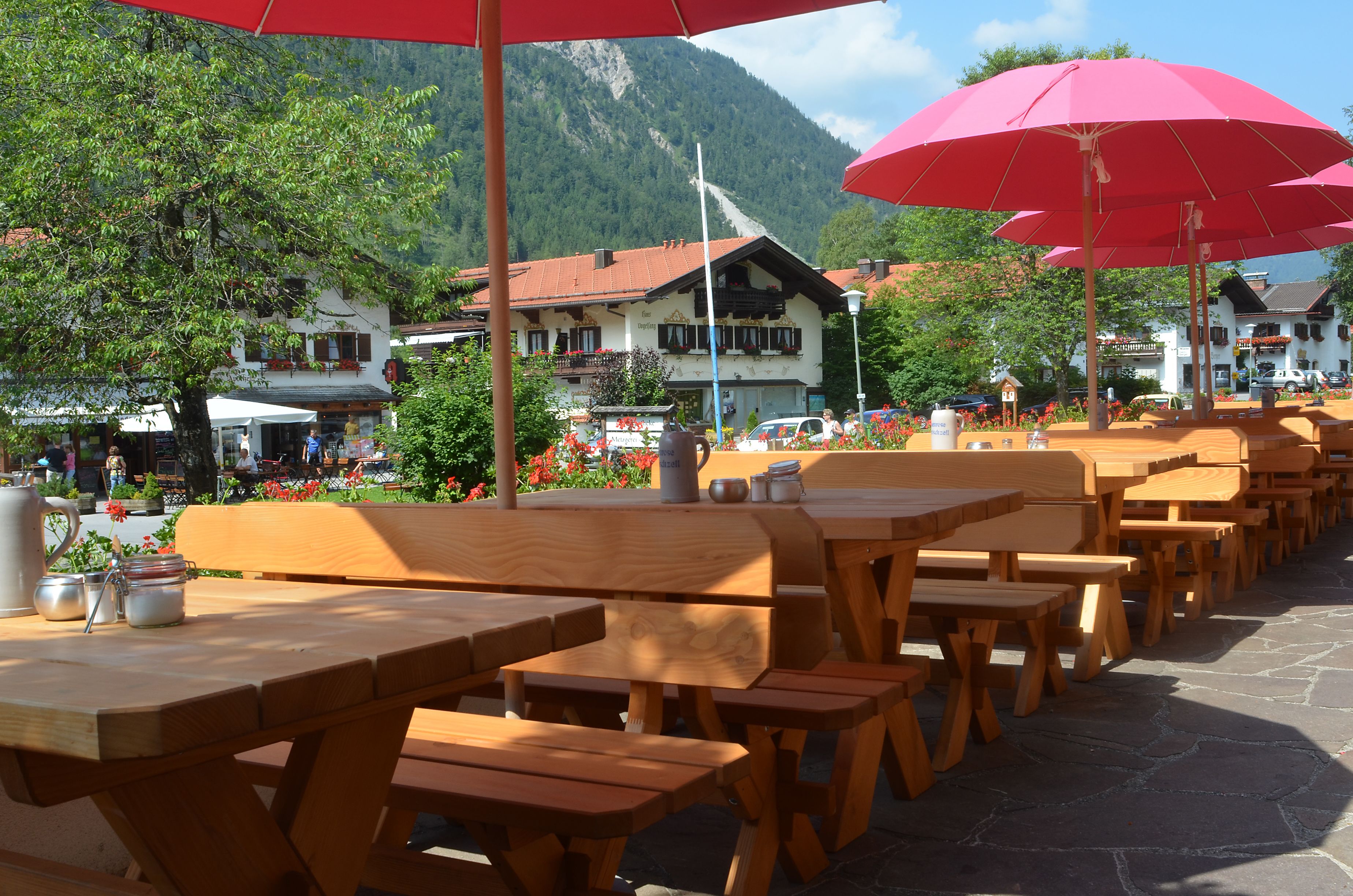 Houten tafels en banken buiten, onder rode parasols, met een berg op de achtergrond en gebouwen verderop.