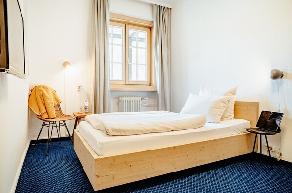 A hotel room with a wooden bed, a chair, a small table with a lamp, and a window with curtains.
