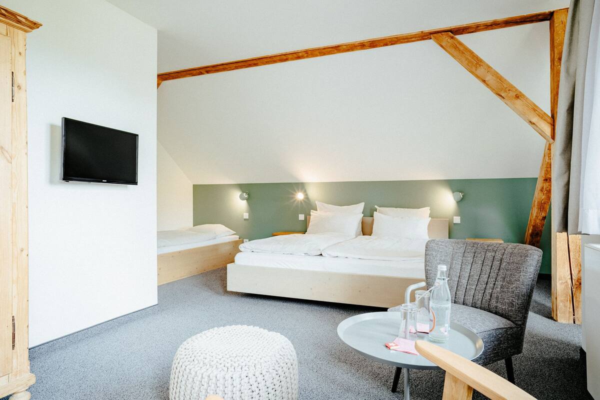 Ein Hotelzimmer mit Bett, Fernseher, Sessel, kleinem Tisch und einem Fenster mit Vorhängen.