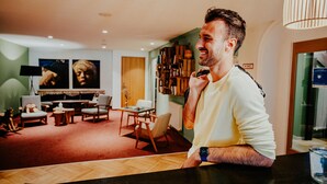 Een man in een informele setting met een modern interieur, inclusief een zithoek met meubels en een boekenkast.