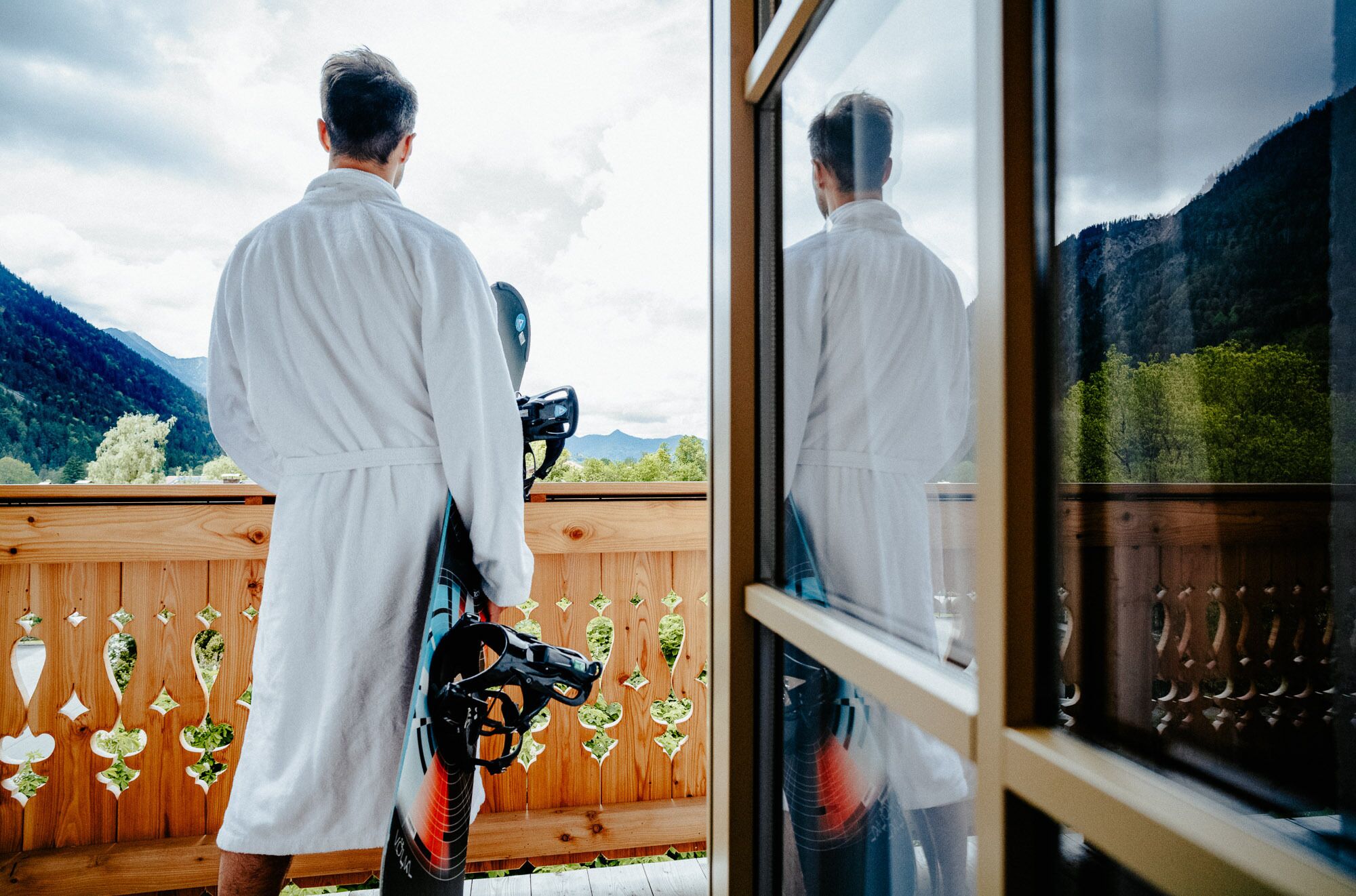 A person in a white robe holding a snowboard on a wooden deck with a scenic mountain view.