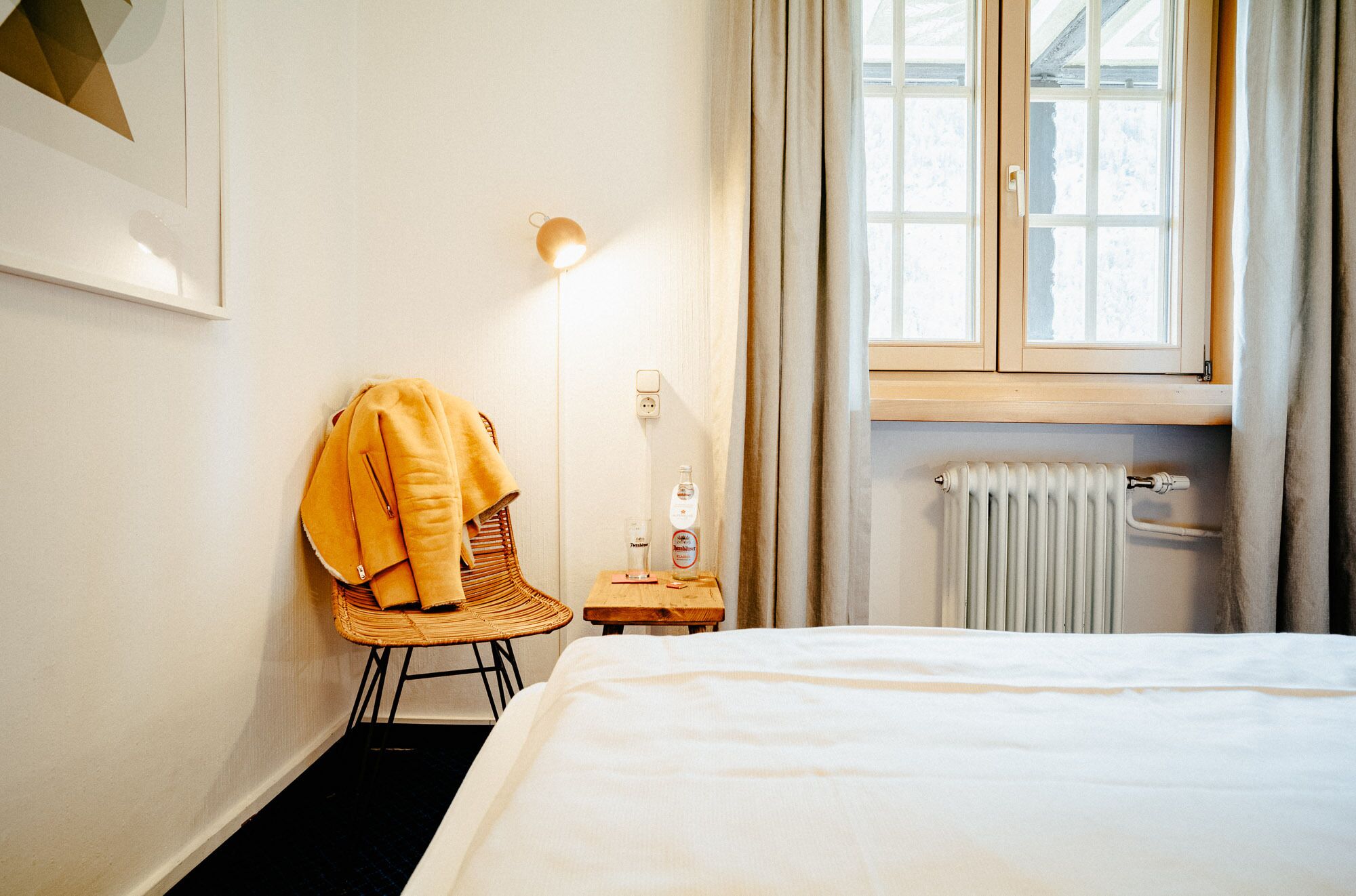 Une chambre d’hôtel avec un lit, une chaise, une table avec une bouteille, une lampe et une fenêtre avec des rideaux.