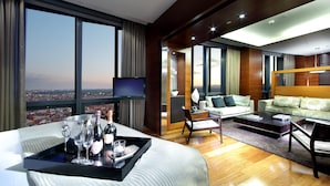 Une chambre d’hôtel moderne dotée d’un grand lit, d’un coin salon avec un canapé et des fauteuils, d’une table à manger et offrant une vue sur la ville grâce à de grandes fenêtres.