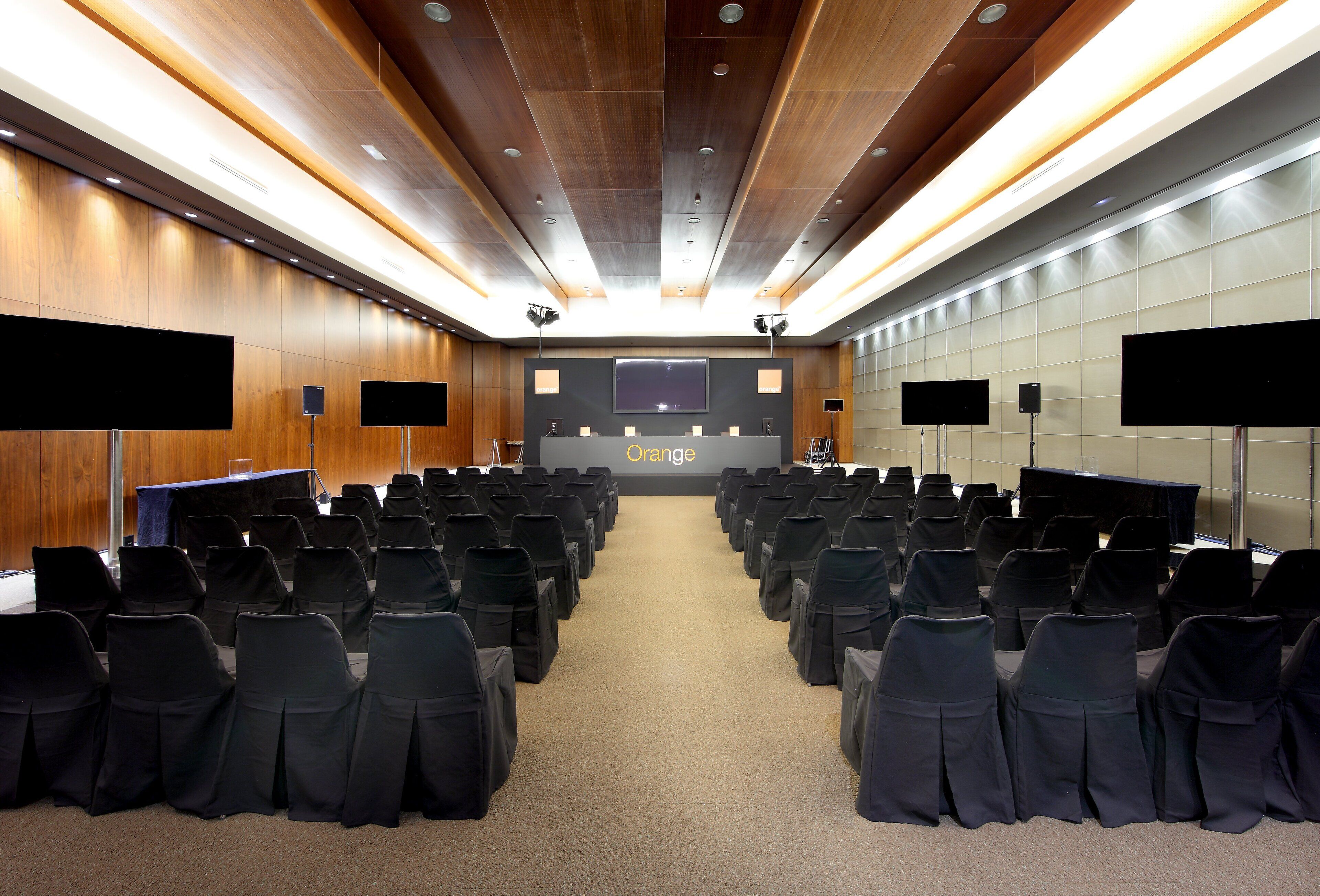 Un salón de conferencias con filas de sillas, un escenario con un atril y una pantalla grande.