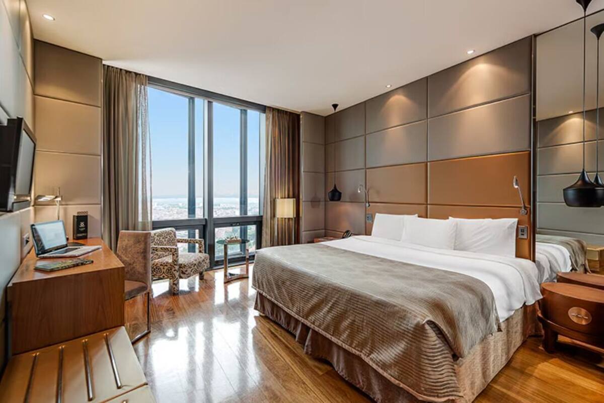 A modern hotel room with a large bed, a desk with a laptop, a chair, and a view of the city through large windows.