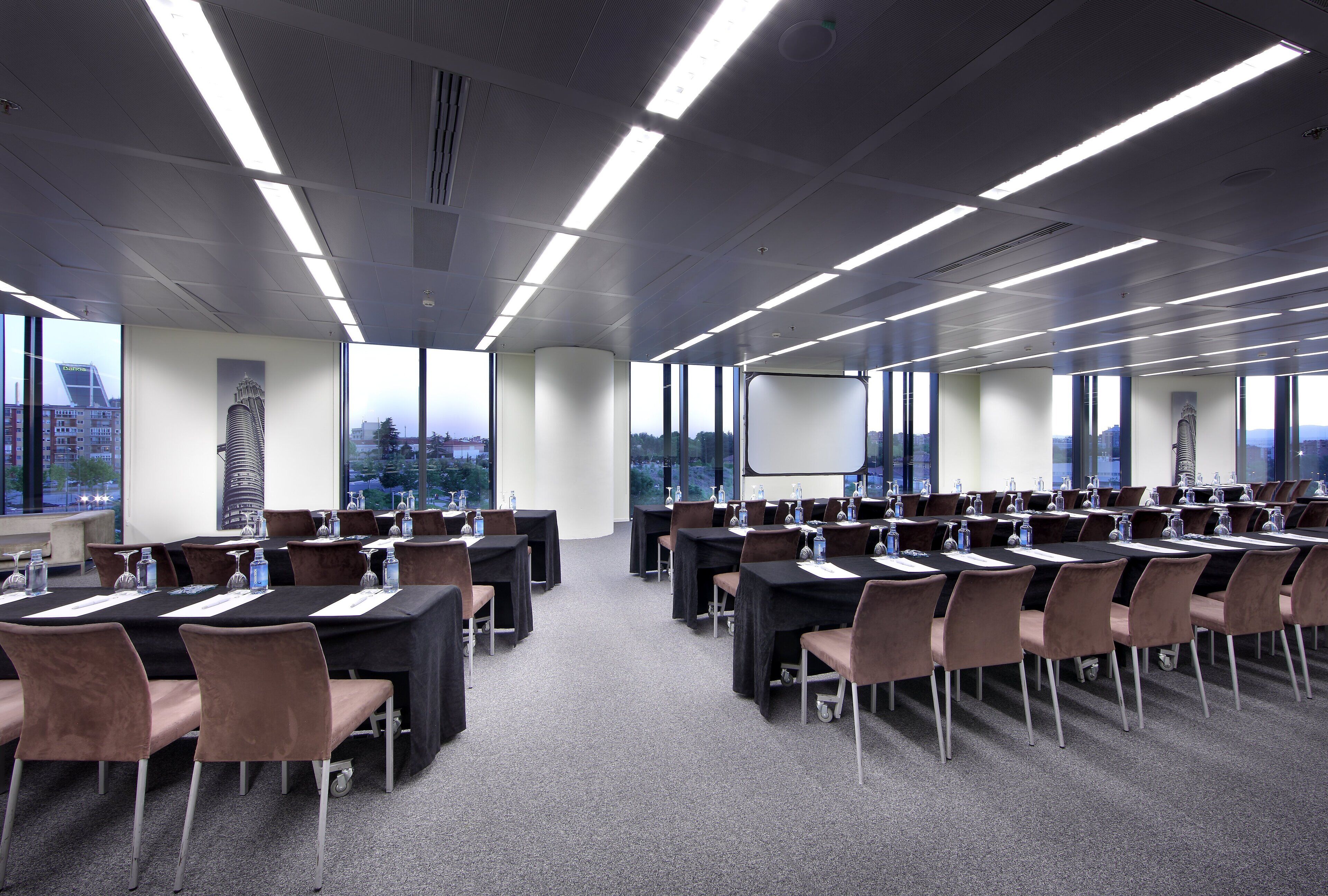 Sala de conferências com várias mesas e cadeiras, grandes janelas e tela de projeção.