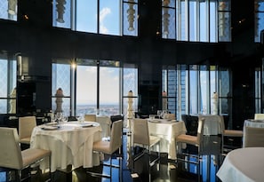 A restaurant with round tables, white tablecloths, and chairs. Large windows offer a view of the cityscape. - Eurostars Madrid Tower (Madrid)