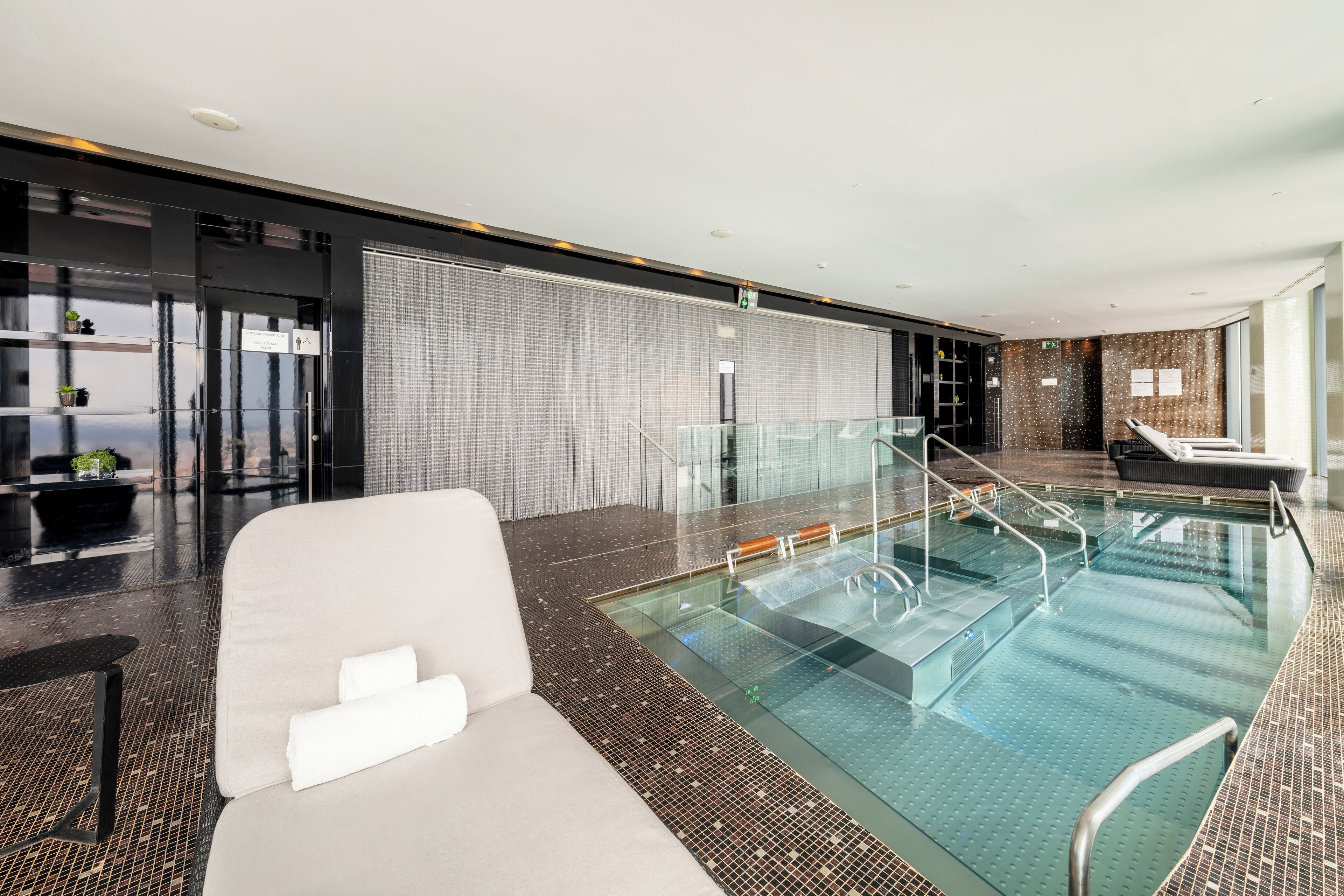 Una piscina interna moderna con ringhiere in vetro, una zona relax con un divano bianco e un muro rivestito di piastrelle con motivo.