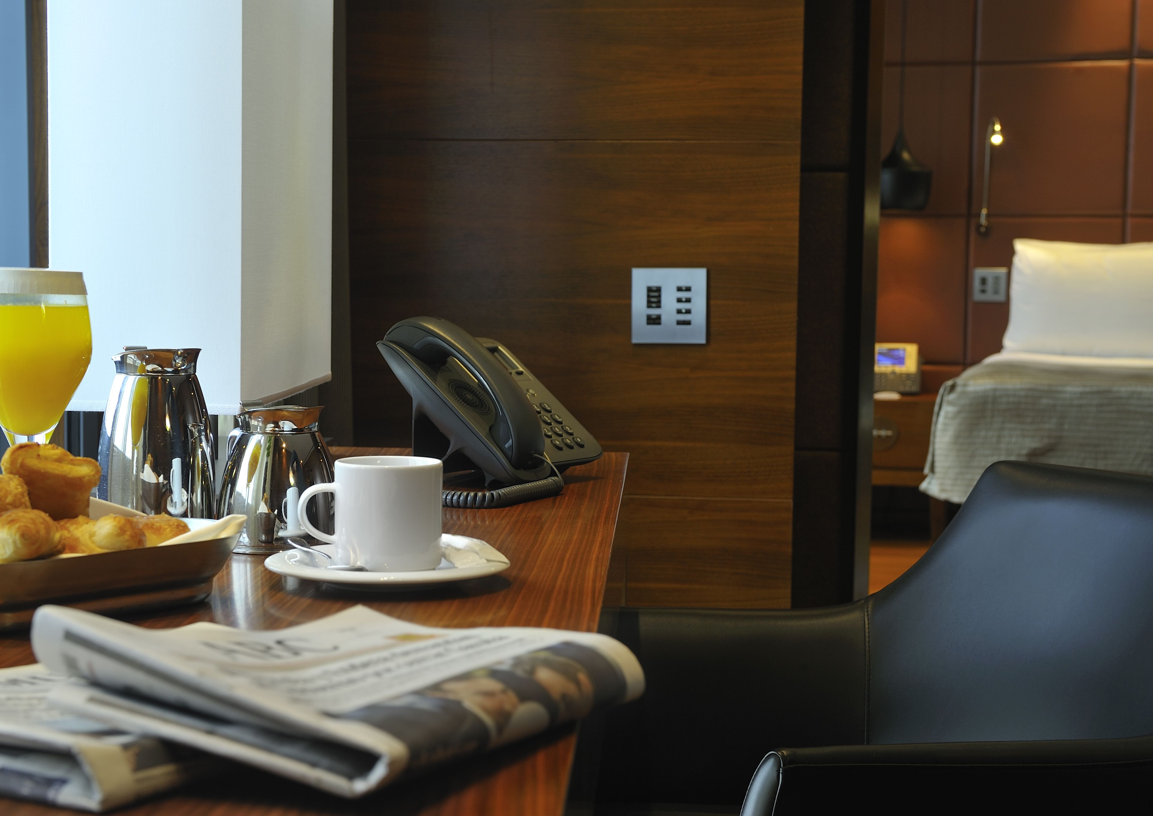 Habitación de hotel con escritorio de madera, teléfono, cafetera, croissants, una taza de café y un periódico.