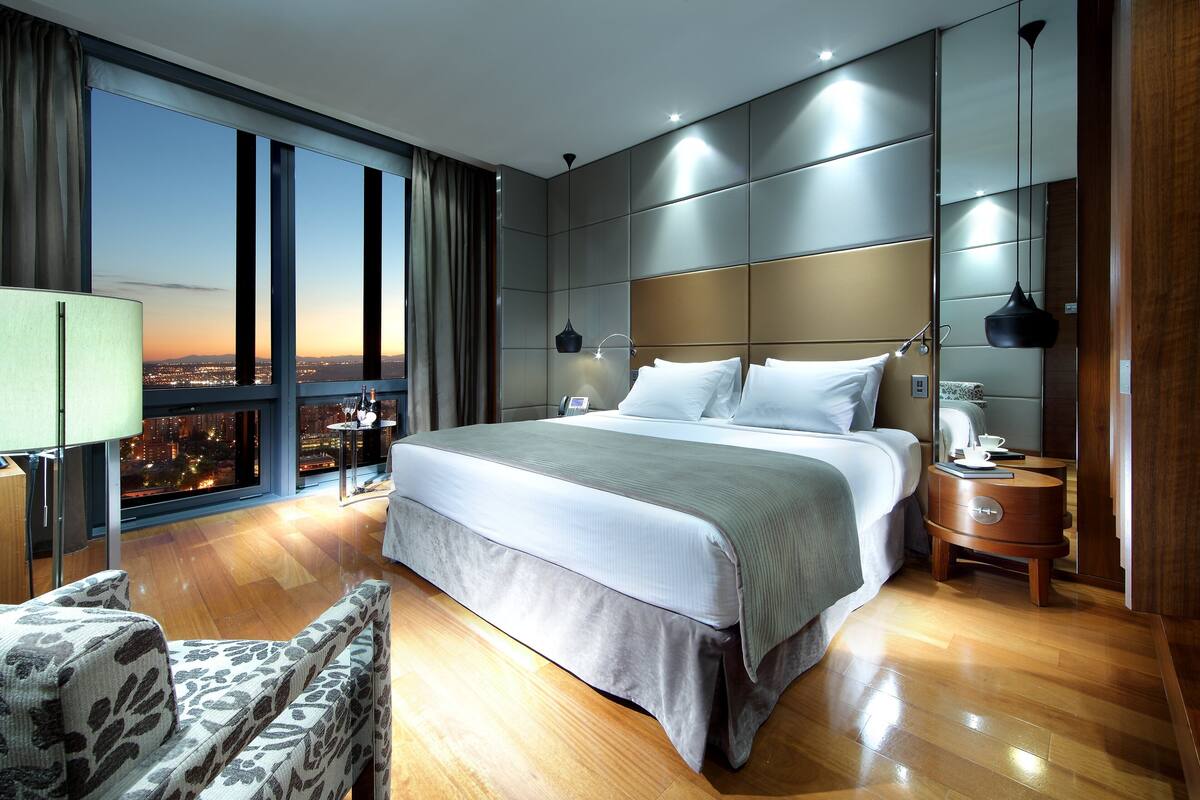 A modern hotel room with a large bed, a view of the city at sunset, a wooden floor, and a nightstand with a lamp.