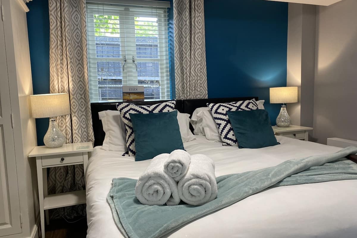 Una cama bien hecha con sábanas blancas, almohadas estampadas en verde agua y blanco, y una manta verde agua. Hay dos mesitas de noche con lámparas y una mesita con una lámpara. La habitación tiene paredes azules y una ventana con cortinas estampadas.