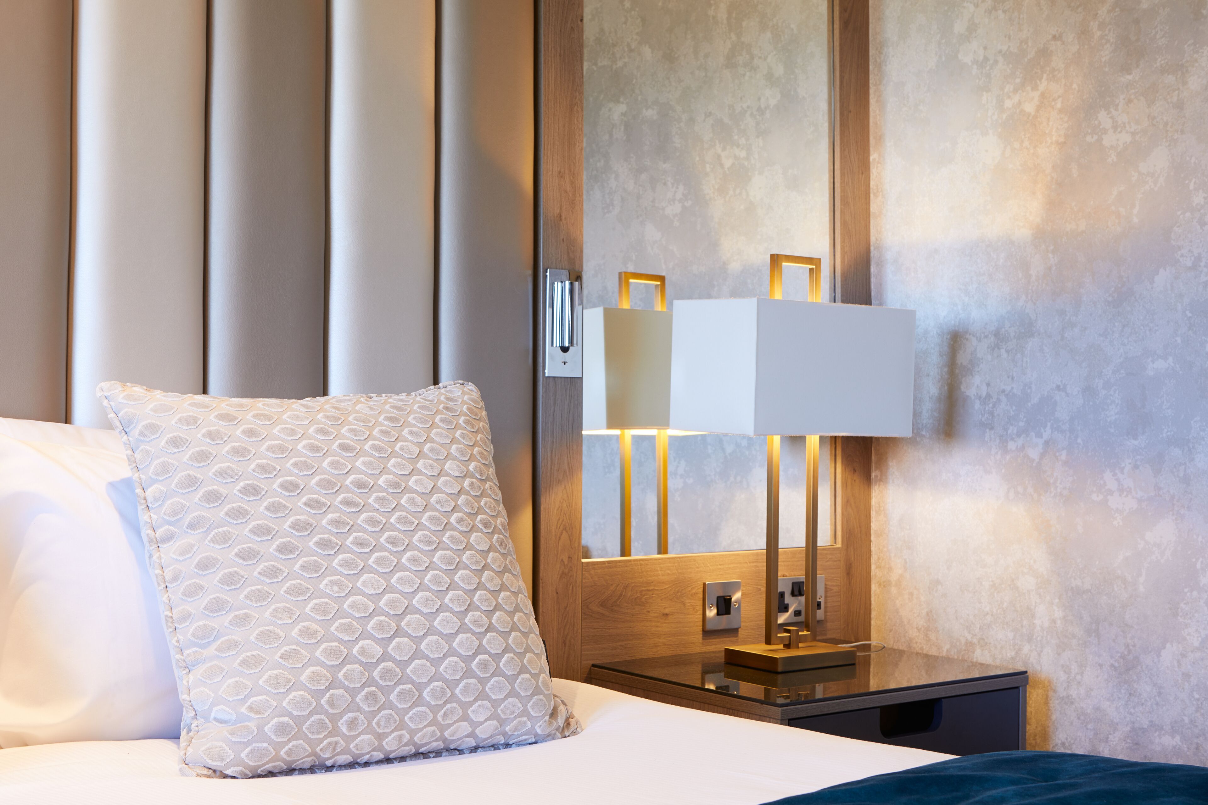 Ein Hotelzimmer mit einem ordentlich bezogenen Bett, einer Nachttischlampe und einem Holennachttisch.