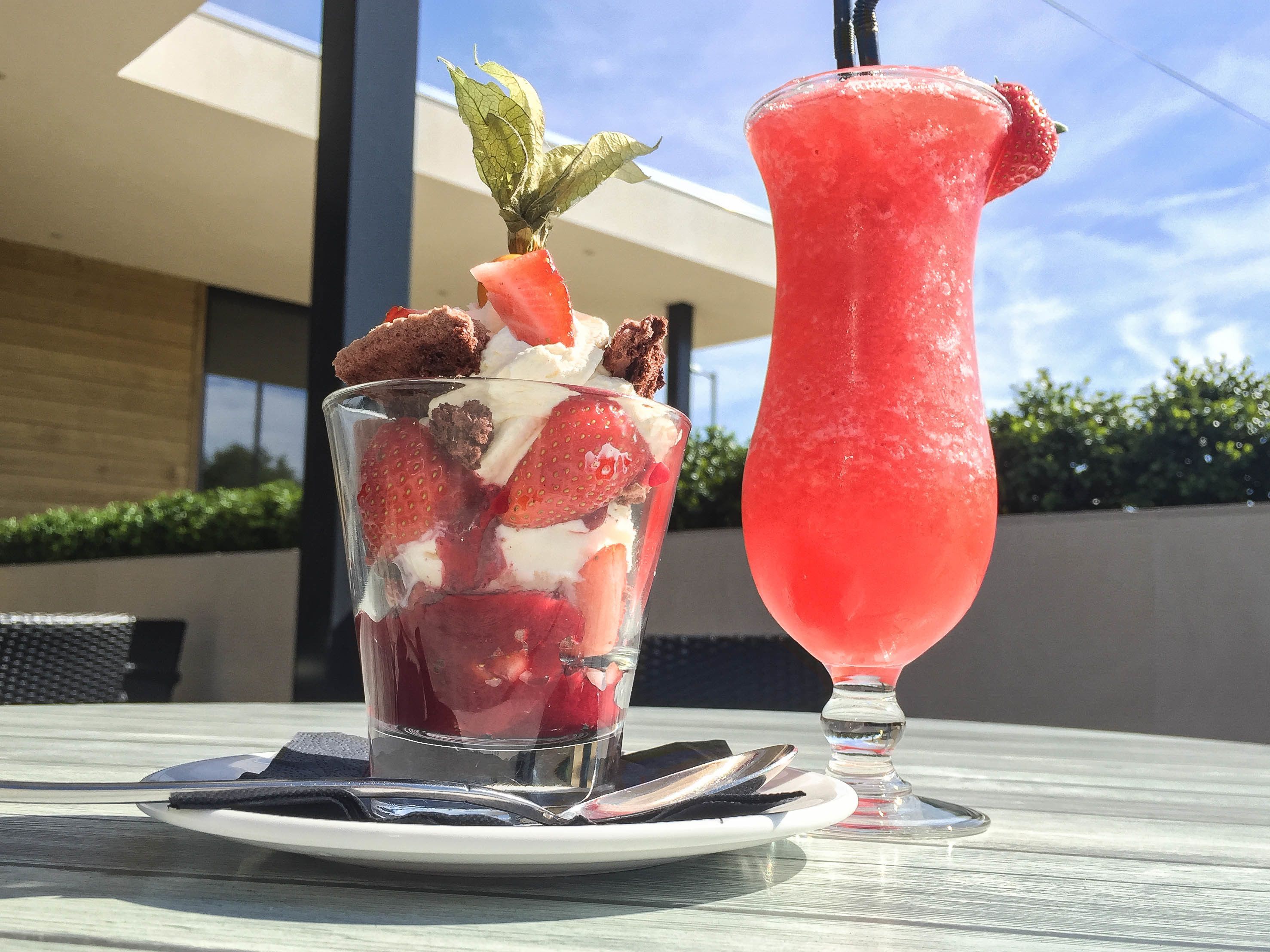 Un postre con fresas y crema batida sobre un plato de vidrio, y un vaso alto de bebida rosa con una pajita sobre una mesa.