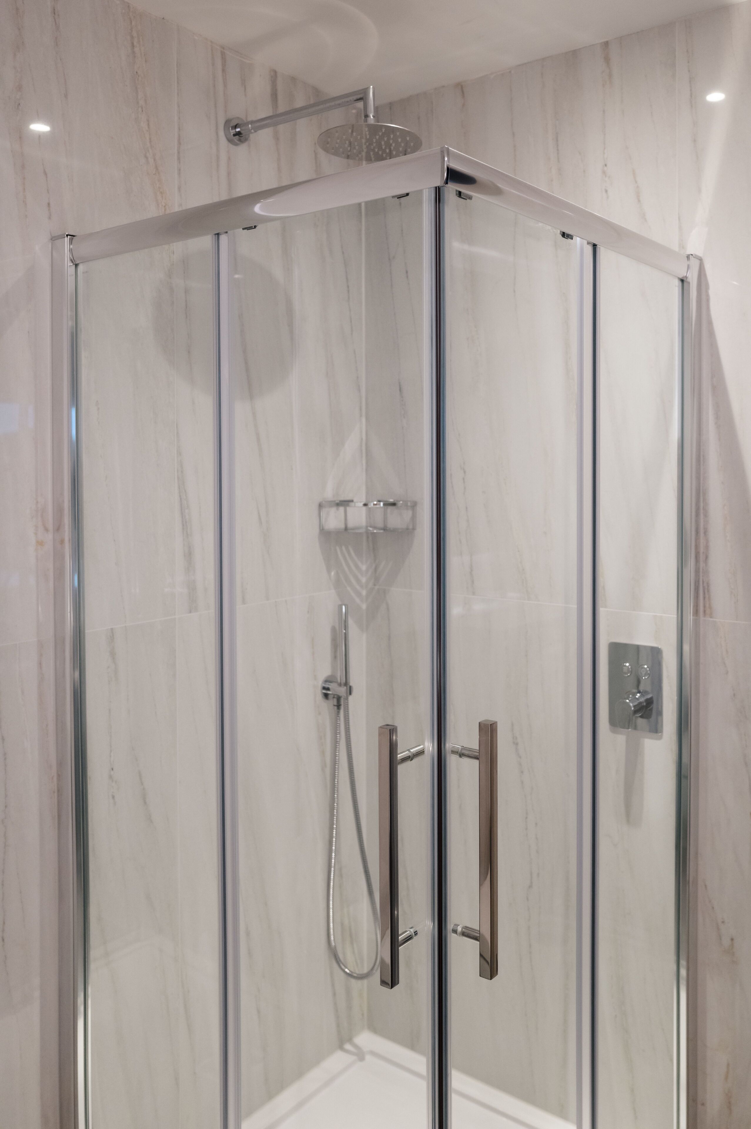 Ducha con mampara de vidrio, cabezal de ducha de mano y panel de control en la pared.